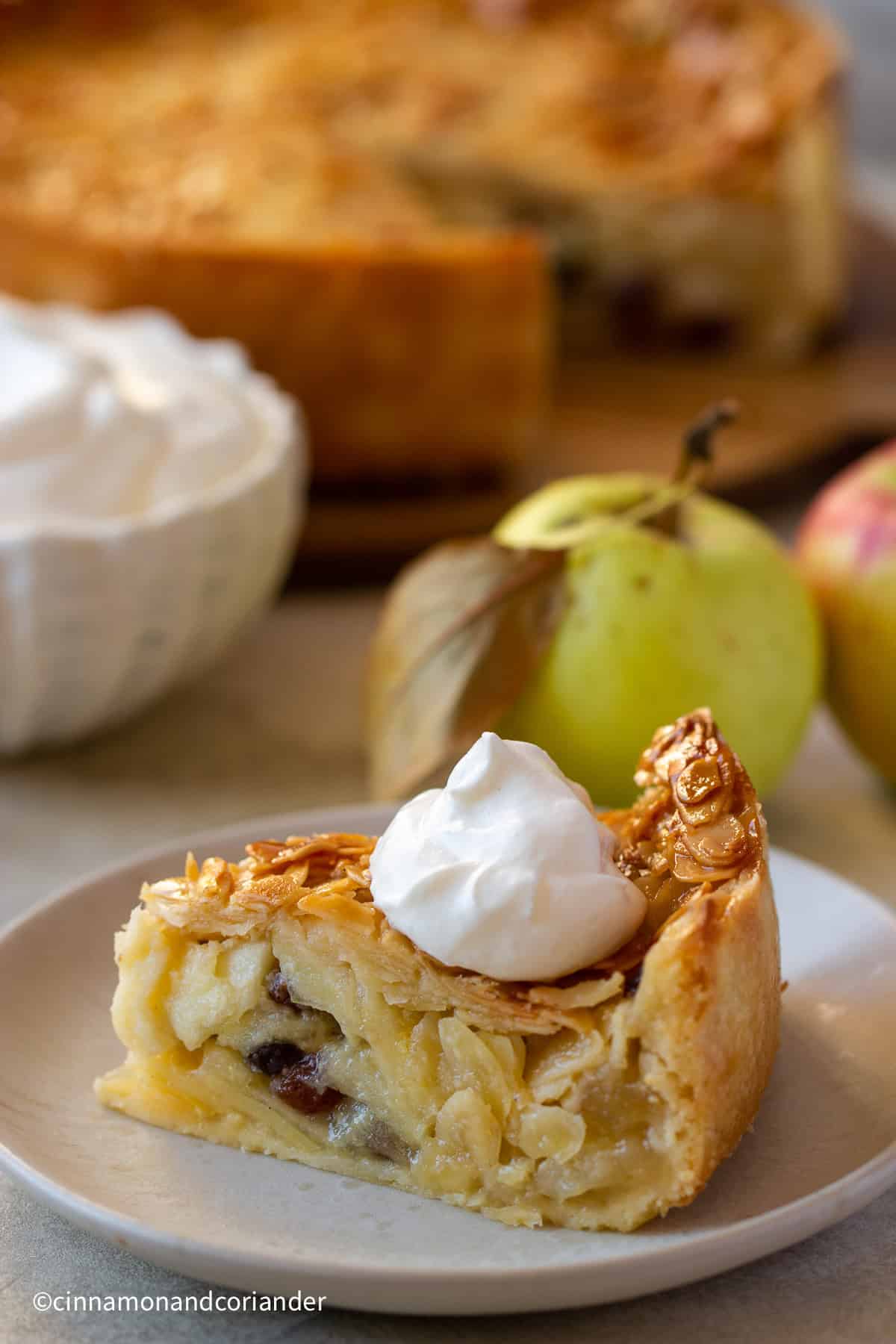ein Stück Florentiner Apfelkuchen mit Mandelkruste mit einem Klecks Sahne serviert mit Äpfeln und einer Sahneschüssel im Hintergrund